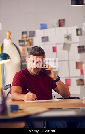 Mature Male Fashion Designer In Wheelchair Talking On Mobile Phone Sitting At Desk Stock Photo