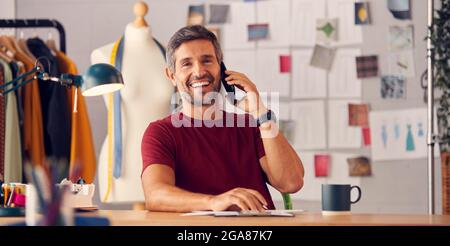 Portrait Of Male Fashion Designer In Studio Working On Sketches Or Documents Making Phone Call Stock Photo