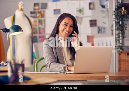 Female Fashion Designer In Studio Working On Sketches Or Documents At Desk Making Phone Call Stock Photo