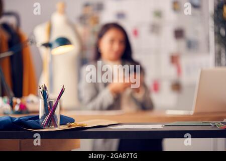 Female Fashion Designer Using Mobile Phone To Browse Internet Sitting At Desk Stock Photo