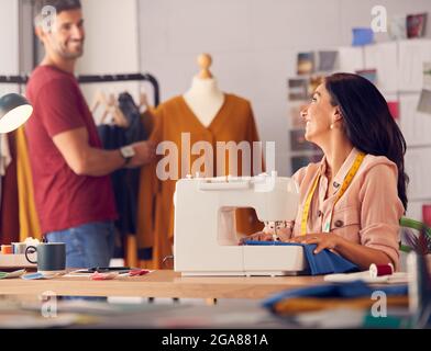 Female Fashion Designer Working On Sewing Machine As Male Colleague Works In Background Stock Photo