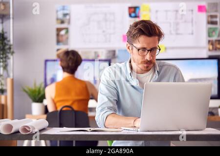 Male And Female Architects In Office Working At Desk On Laptop And Desktop Computers Stock Photo