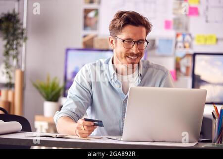 Male Architect In Office Working At Desk Making Online Purchase Using Credit Card On Laptop Stock Photo