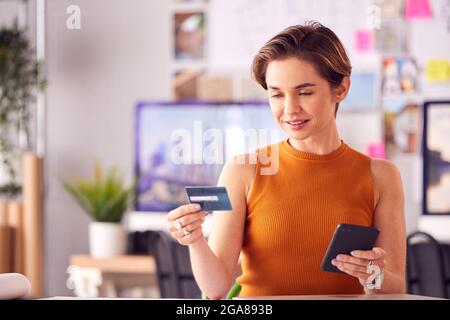 Female Architect In Office Working At Desk Making Online Purchase Using Credit Card On Mobile Phone Stock Photo