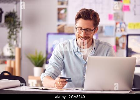 Male Architect In Office Working At Desk Making Online Purchase Using Credit Card On Laptop Stock Photo