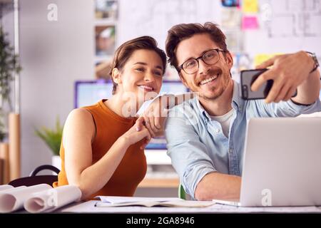 Male And Female Architects In Office Sitting At Desk Posing For Selfie Together Stock Photo