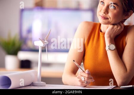 Female Architect In Office Working On Drawings For Renewable Energy Project With Model Wind Turbine Stock Photo