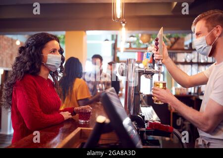 Male Bartender Wearing Face Mask Serving Female Customer With Drink During Health Pandemic Stock Photo