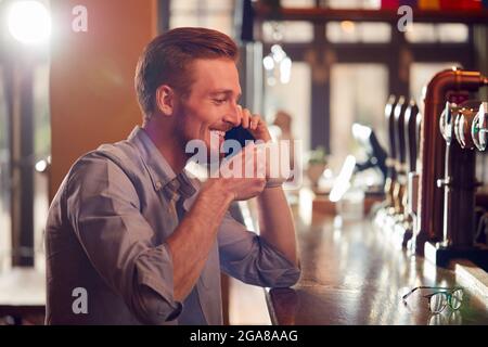 Male Owner Or Manager Of Bar Sitting At Counter Drinking Coffee And Talking On Mobile Phone Stock Photo