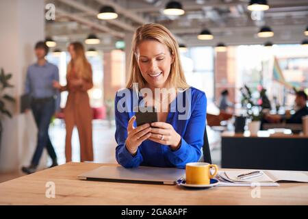 Young Businesswoman Sitting At Desk Using Mobile Phone In Modern Open Plan Office Stock Photo