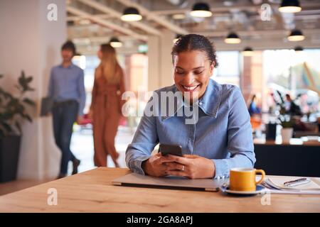 Young Businesswoman Sitting At Desk Using Mobile Phone In Modern Open Plan Office Stock Photo