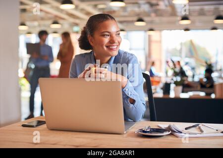 Young Businesswoman With Laptop Sitting At Desk Having Coffee Break In Modern Open Plan Office Stock Photo