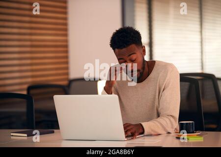 Tired Young Businessman Working Late Sitting At Desk With Laptop In Modern Open Plan Office Stock Photo
