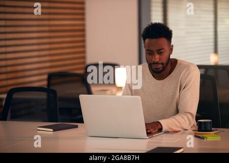 Young Businessman Working Late Sitting At Desk With Laptop In Modern Open Plan Office Stock Photo