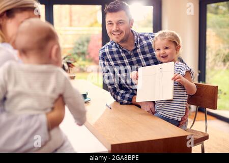 Family Sitting Around Kitchen Counter With Daughter Showing Parent Picture She Has Drawn Stock Photo