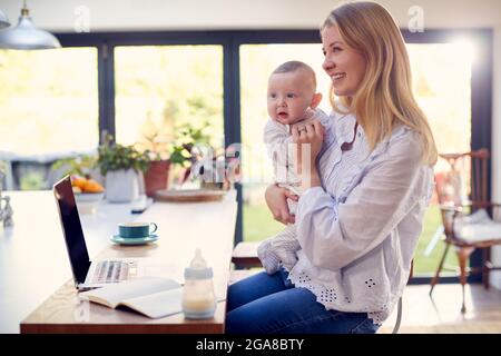 Working Mother Using Laptop At Home Whilst Holding Smiling Baby Son On Knee Stock Photo