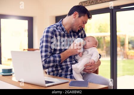 Working Father Using Laptop At Home Whilst Feeding Baby Son On Knee With Bottle Stock Photo