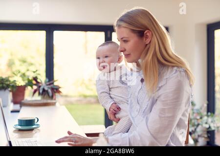 Working Mother Using Laptop At Home Whilst Holding Smiling Baby Son On Knee Stock Photo