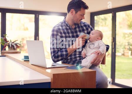 Working Father Using Laptop At Home Whilst Feeding Baby Son On Knee With Bottle Stock Photo