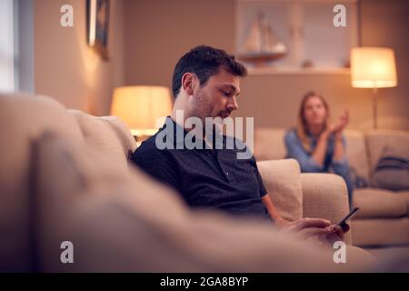 Evening Shot Of Couple On Sofa At Home With Man Looking At Mobile Phone And Unhappy Woman Behind Stock Photo