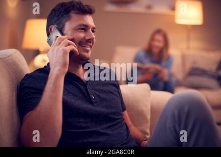 Evening Shot Of Couple Relaxing On Sofa At Home As Man Talks On Mobile Phone And Woman Reads Book Stock Photo