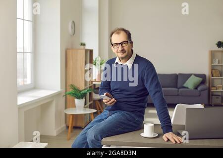 Mature businessman with phone sitting on table with laptop in home office Stock Photo
