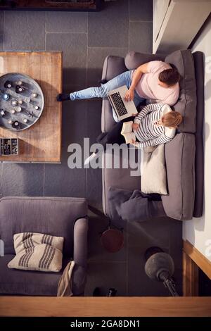 Overhead View Of Couple Relaxing On Lounge Sofa At Home Reading Book And Using Laptop Stock Photo