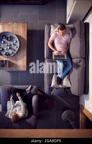 Overhead View Of Couple Relaxing On Lounge Sofa At Home And Watching TV And Working On Laptop Stock Photo