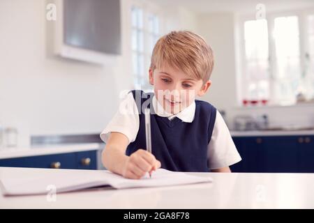 Boy At Home Wearing School Uniform Doing Homework On Kitchen Counter Stock Photo