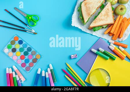 Notebooks, pens, stationery and a breakfast of sandwich, apples and carrots on a blue background. Top view, copy space. Back to school. Stock Photo