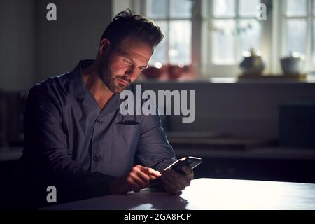 Man Wearing Pyjamas Sitting In Kitchen At Night Using Mobile Phone Stock Photo