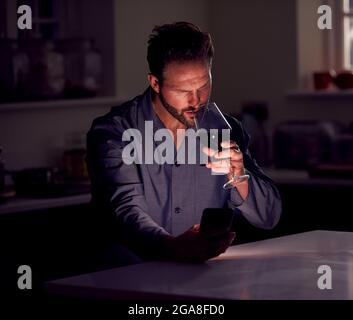 Man Wearing Pyjamas Sitting In Kitchen With Glass Of Wine At Night Using Mobile Phone Stock Photo