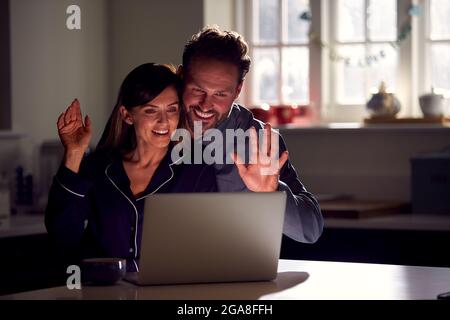 Couple Wearing Pyjamas Sitting In Kitchen At Night Using Laptop For Video Call Stock Photo