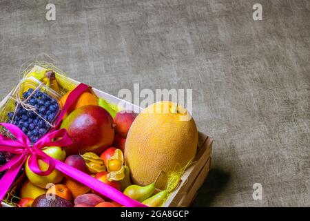 Ripe fruits and berries are beautifully stacked in wooden box tied with pink ribbon. Dark background. Food delivery, gift fruit set. Concept of health Stock Photo