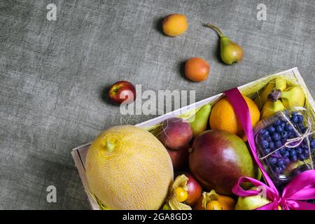 Ripe fruits and berries are beautifully stacked in wooden box tied with pink ribbon. Dark background. Food delivery, gift fruit set. Concept of health Stock Photo