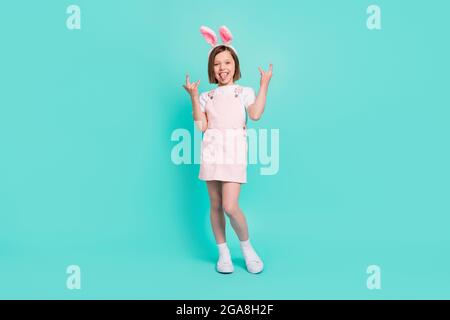 Photo of impressed funky school girl wear pink overall bunny ears smiling showing tongue hard rock sign isolated teal color background Stock Photo