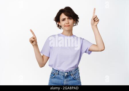 Image of skeptical female model grimacing, frowning and pointing sideways, two doubtful things, express dislike, complaining, standing against white Stock Photo