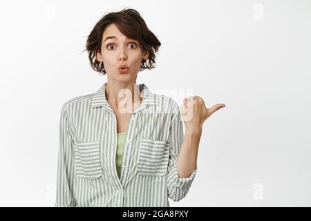 Excited brunette girl looks interested, points right, asks question about banner or product you place on empty copy space, stands against white Stock Photo