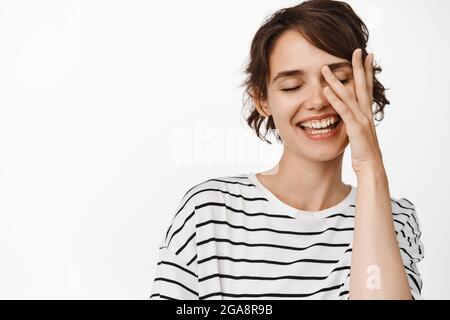 Beautiful young woman with perfect clean, healthy skin, no makeup, smiling and gently touching face with one hand, standing against white background Stock Photo