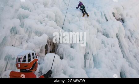 Man standing and controlling a safety top rope while female with ice climbing equipment, climbing on a frozen waterfall Stock Photo