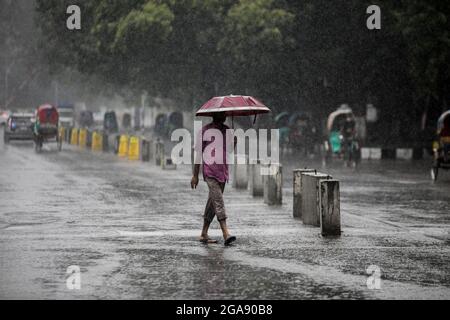 Dhaka, Bangladesh. 29th July, 2021. A man seen holding an umbrella while walking along an empty and deserted road during a rainy day amid the covid-19 lockdown.The country went into a strict lockdown and Heavy monsoon downpour caused extreme water logging in most areas of Dhaka city. Roads were submerged making travel slow and dangerous. (Credit Image: © Sazzad Hossain/SOPA Images via ZUMA Press Wire) Stock Photo