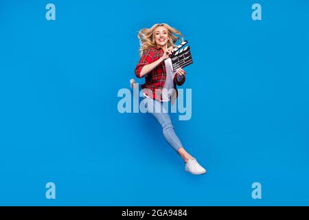 Photo of funky sweet young lady wear plaid shirt smiling humping high holding clapperboard isolated blue color background Stock Photo