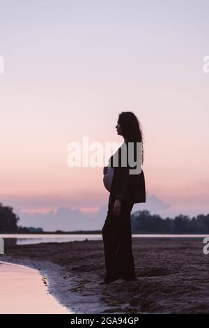 Silhouette of pregnant woman on beach. Beautiful young brunette with long curly hair stands on sand on beach and enjoys nature and setting sun