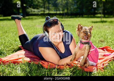 Overweight woman in wireless headphones lies in park with dog and smiles. Stock Photo