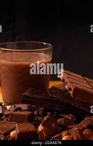 Set of chocolate milkshake glass and chocolate bars on a pile of chocolate chips, cocoa powder and mixed nuts placed in front of a black background Stock Photo