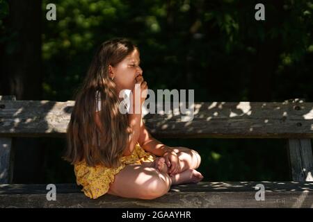 Cute funny Caucasian Girl sitting on the bench and eating ice cream . Smiling and dreaming kid. Summer time. Happy childhood. Stock Photo