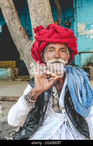 Portrait of Indian male villager in a red turban and tribal jewelry smoking a cigarette in a village in Rajasthan, India Stock Photo