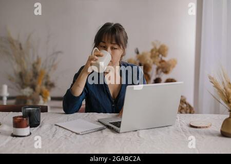 Busy focused asian woman freelancer drinking tea or coffee while working remotely on laptop from home Stock Photo
