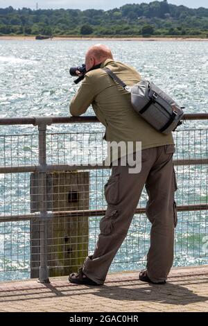 older photographer leaning on railings taking a picture of the sea with a digital camera and holding a camera bag Stock Photo