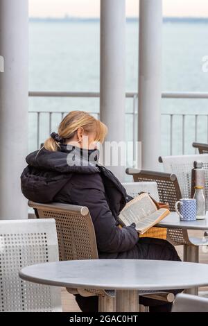 young female sitting outdoors reading a book, lady sitting at cafe reading a book, young woman reading a book out of doors, reader with book engrossed Stock Photo
